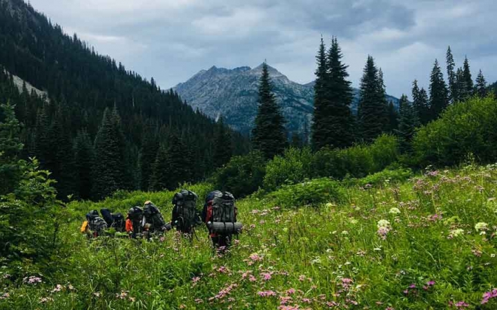 a group of students carrying backpacks hike through a green mountain meadow with mountains in the background